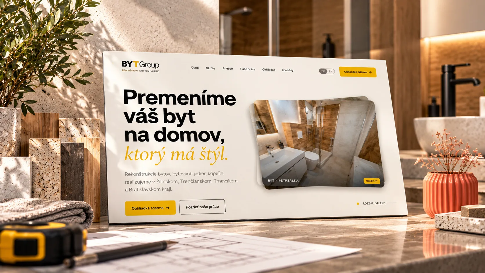 Úvodná stránka webu BYTGroup na obrazovke medzi vzorkami kameňa a dreva, metrom, výkresom a koralovou vázou